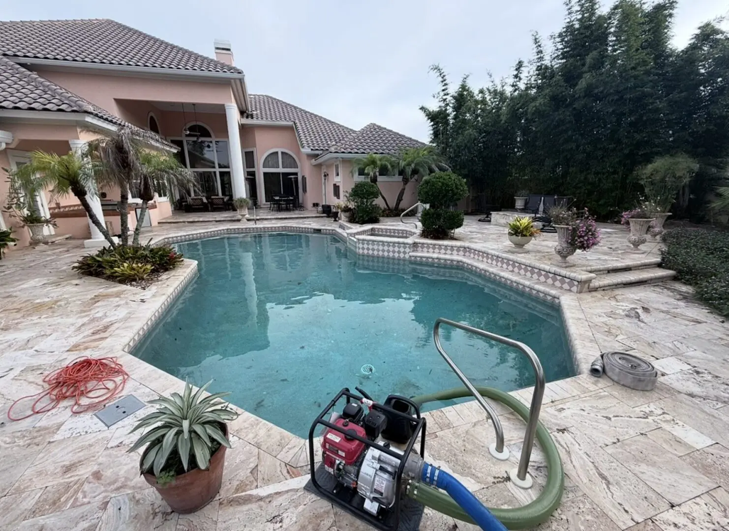 Swimming pool with a hose attached to it in front of a house, surrounded by lush plants and trees