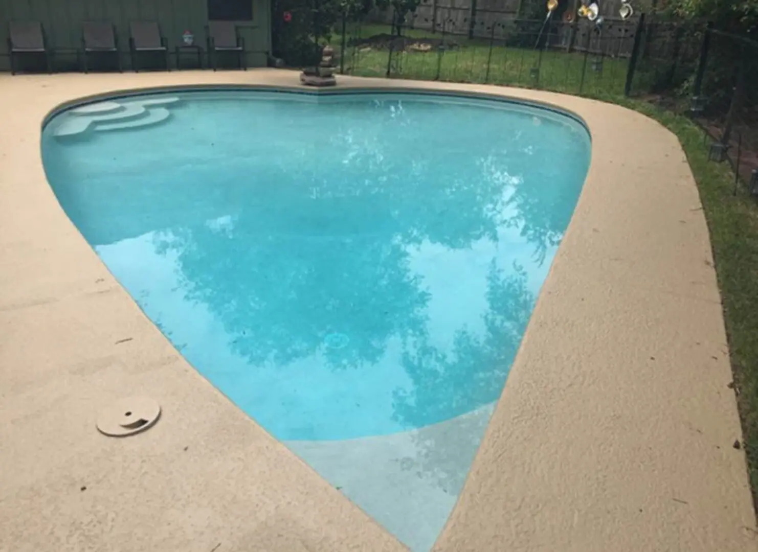 Heart-shaped swimming pool in a backyard surrounded by a fence, with a house in the background, chairs, trees, and grass
