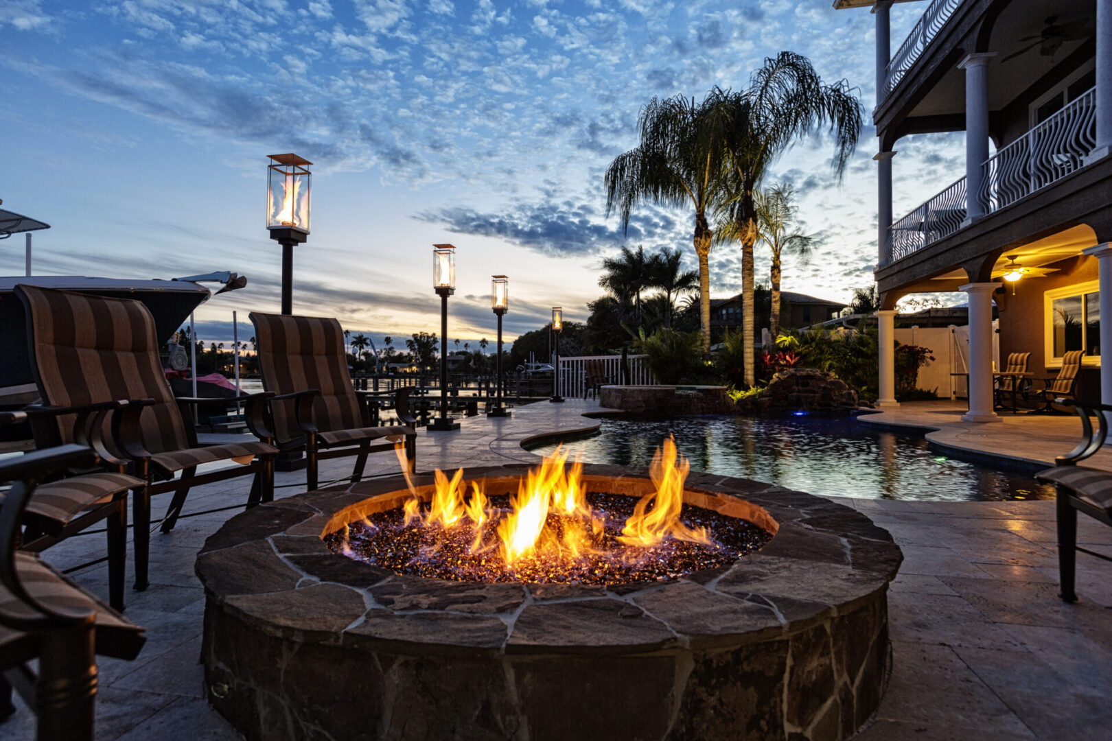 Fire pit sitting on top of a patio next to a swimming pool, surrounded by chairs, light poles, plants, trees