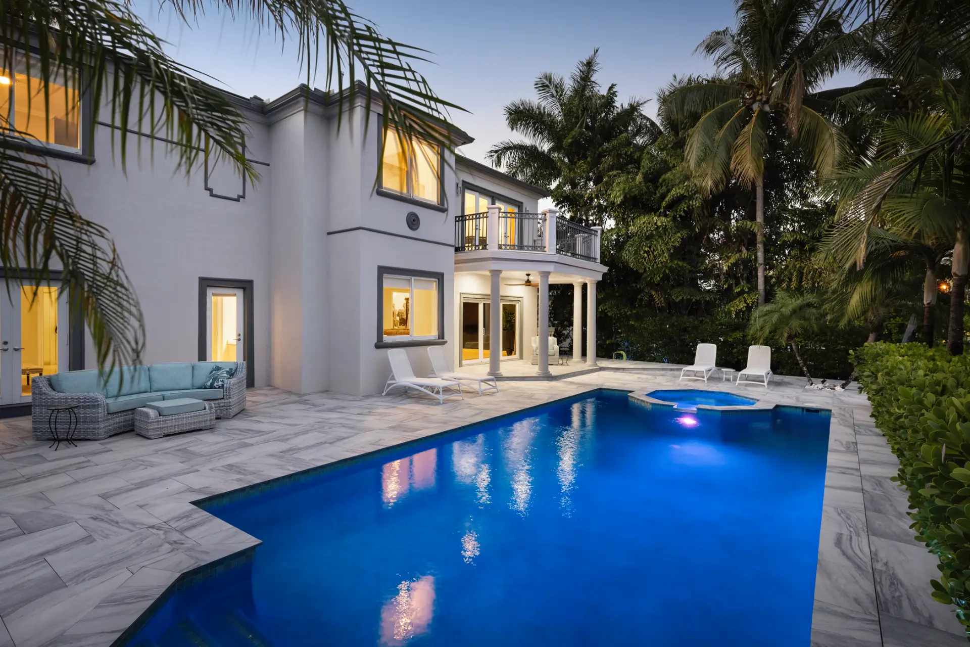 Large white house with a swimming pool in front of it, surrounded by lush green plants and trees
