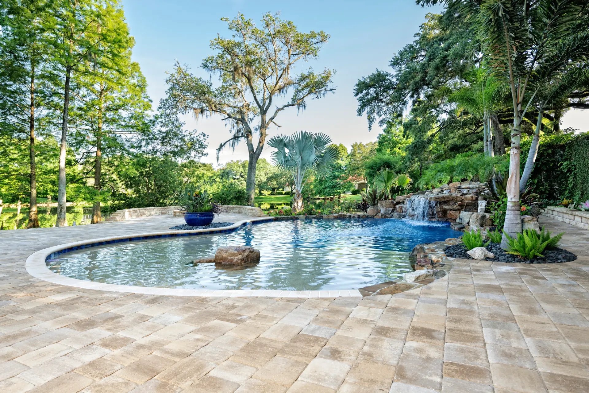 Swimming pool with a waterfall in the middle of it surrounded by lush greenery, including trees, plants, and rocks