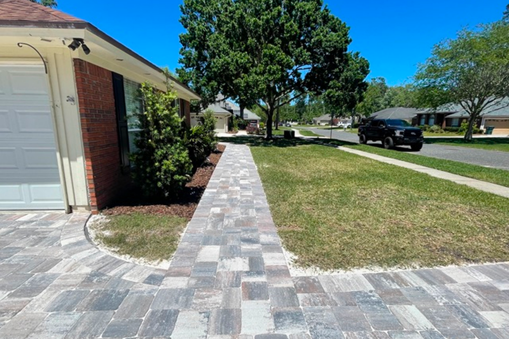 Brick walkway to a house with a car parked in front of it, surrounded by lush green grass, plants