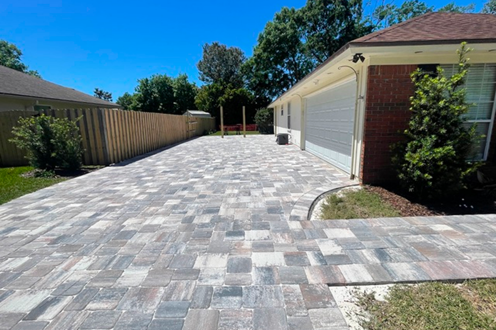 Brick driveway with a brick paver walkway in front of a house, surrounded by lush green grass, plants, and trees