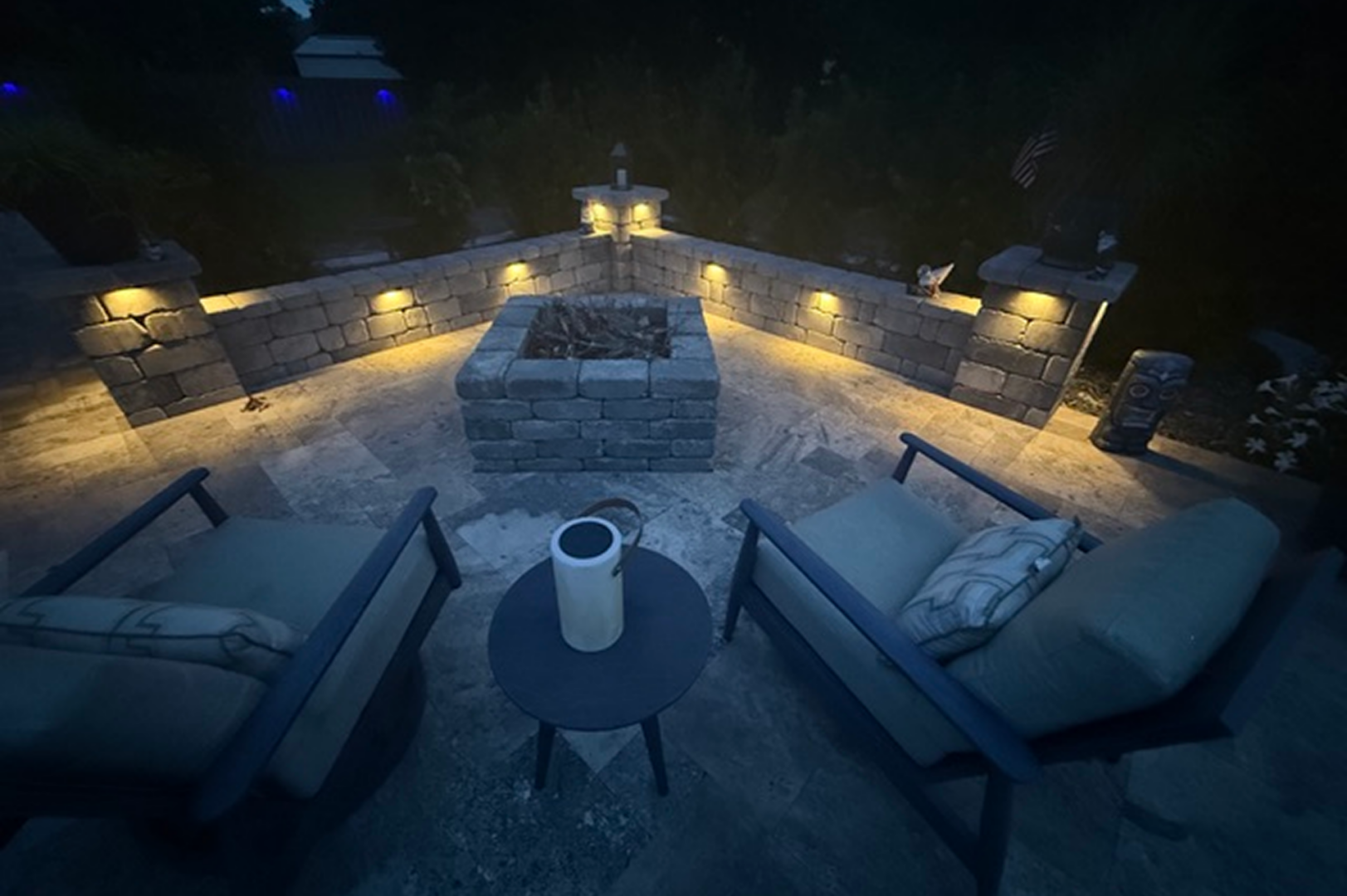 Patio with two chairs and a fire pit at night, surrounded by a wall with lights