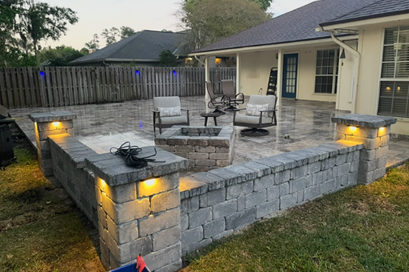 Brick patio with a fire pit in the middle of it, surrounded by chairs and lights