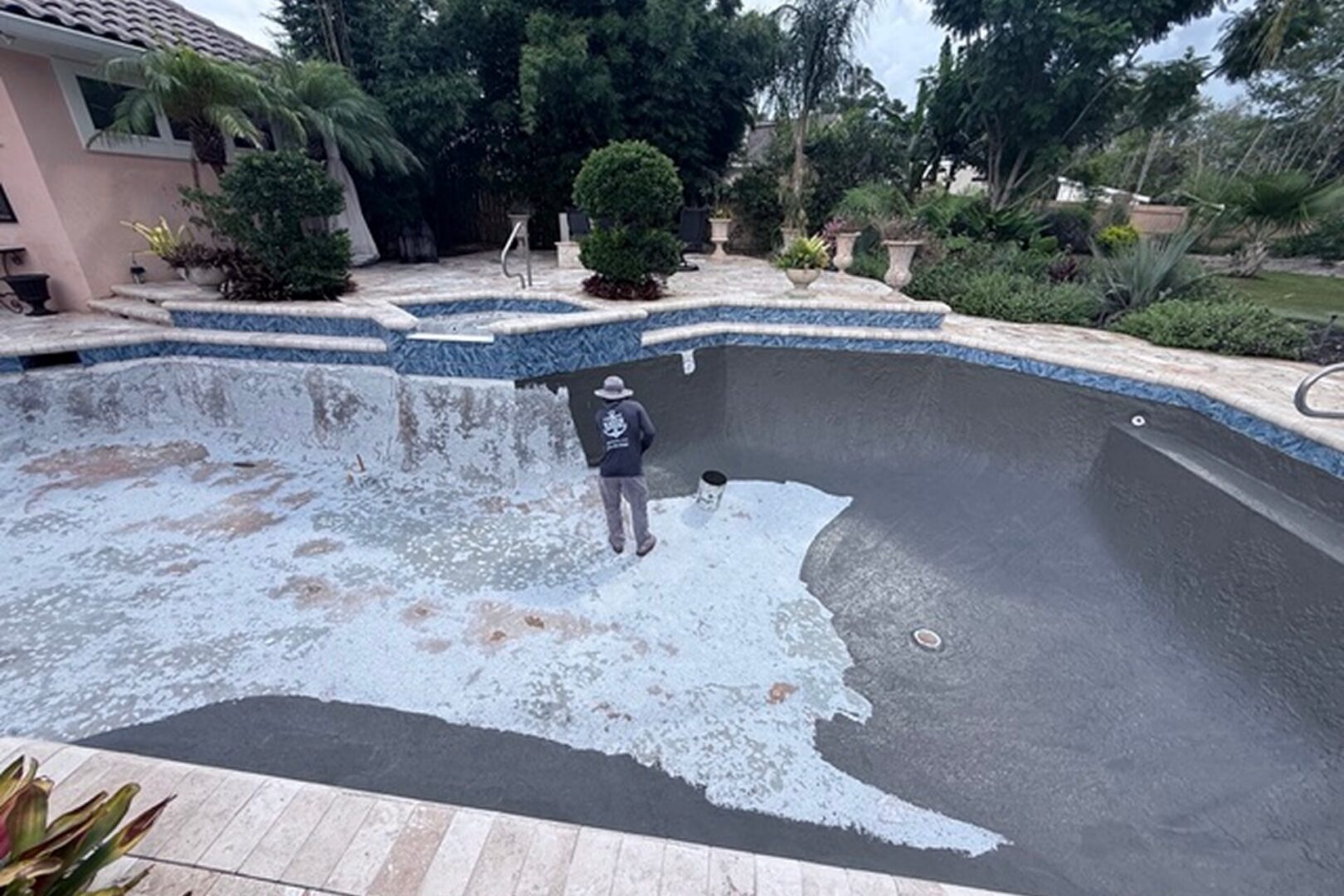 Man standing in the middle of a swimming pool surrounded by plants, trees, and a house in the background